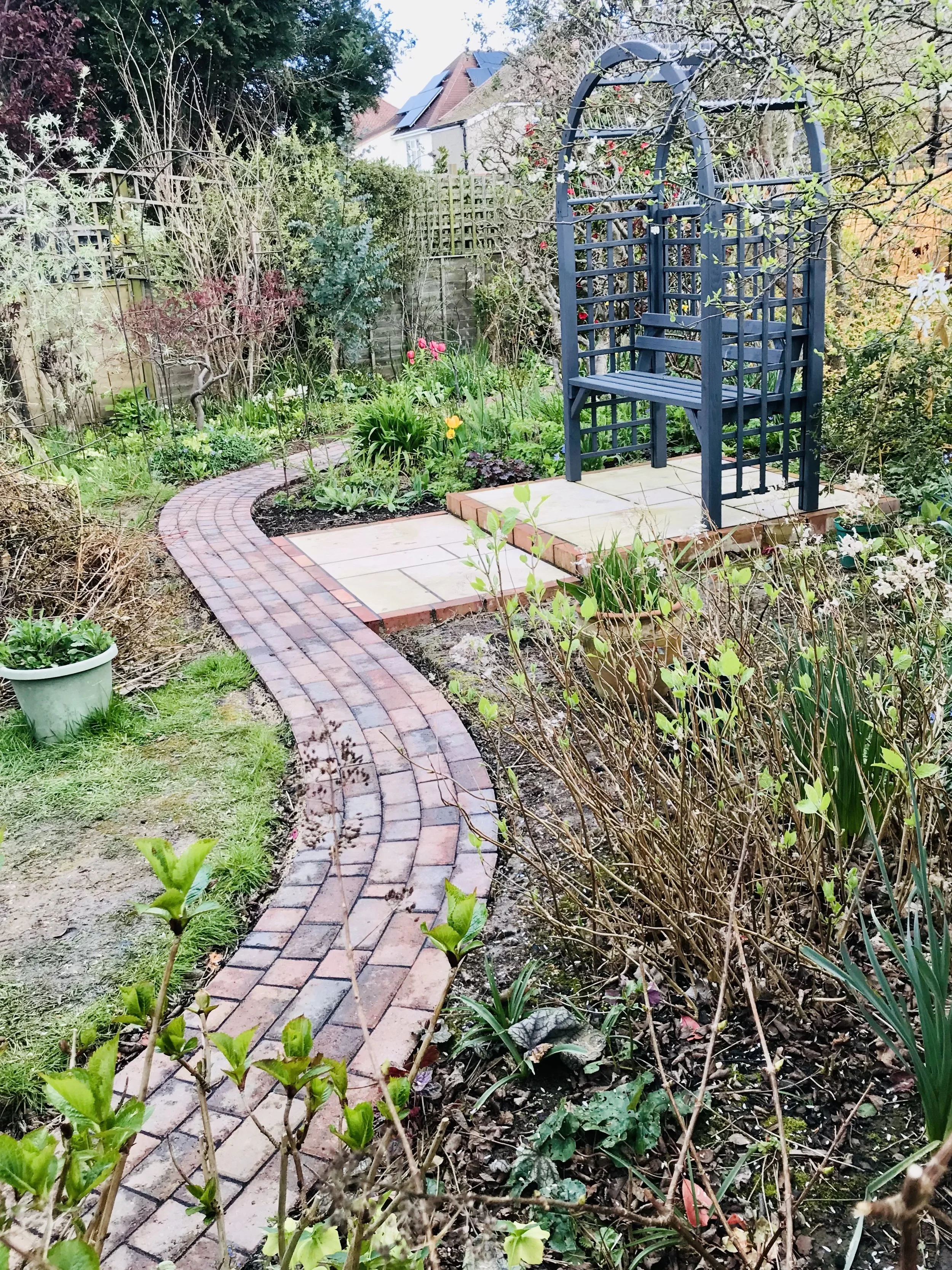 A garden with a curved brick pathway leading to a wooden platform with a blue garden bench. There are various plants and flowers surrounding the pathway and bench, and a brick-backed fence in the background.