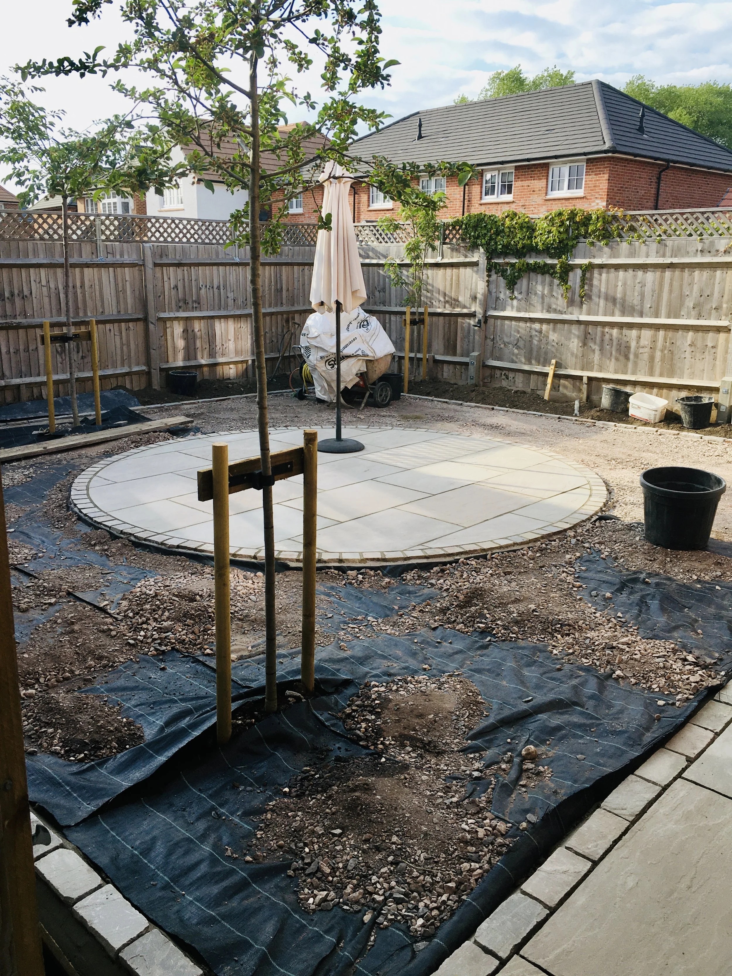 Backyard patio under construction with some paving stones laid in a circular pattern, young trees staked for support, and construction materials and tools around the area.