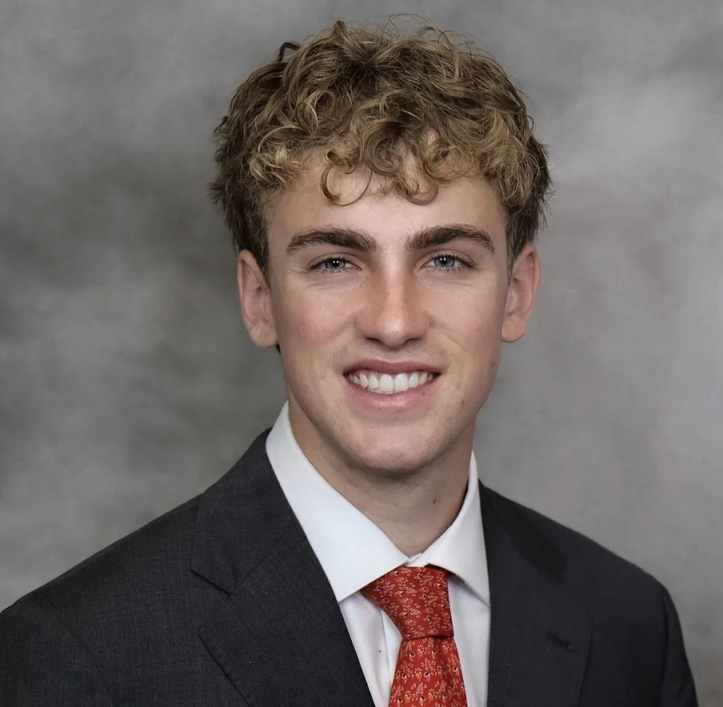 Portrait of a young man with curly blond hair, blue eyes, wearing a dark suit, white shirt, and red patterned tie, smiling against a gray background.