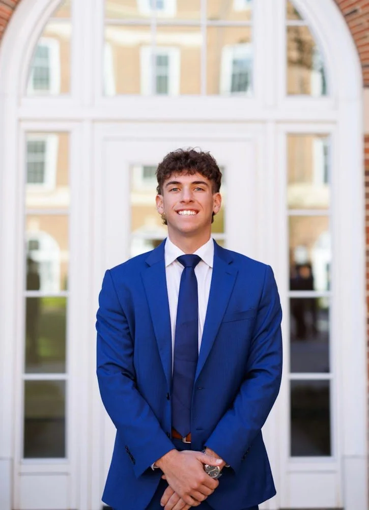 Young man in a blue suit and tie smiling in front of a large window with buildings reflected in the glass.