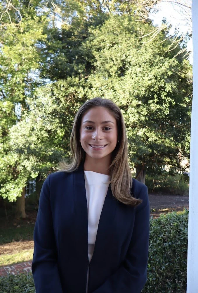A young woman with long brown hair smiling outdoors, wearing a navy blazer over a white top, standing in front of lush green trees.