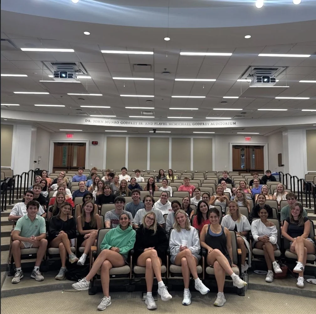 A large group of students sitting in an auditorium with beige seats, facing forward and smiling for the photo.