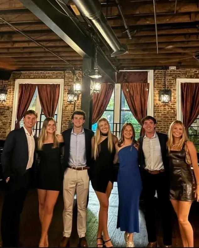 Group of seven young adults dressed in semi-formal attire, standing inside a brick-walled venue with large windows, red curtains, and hanging lights.