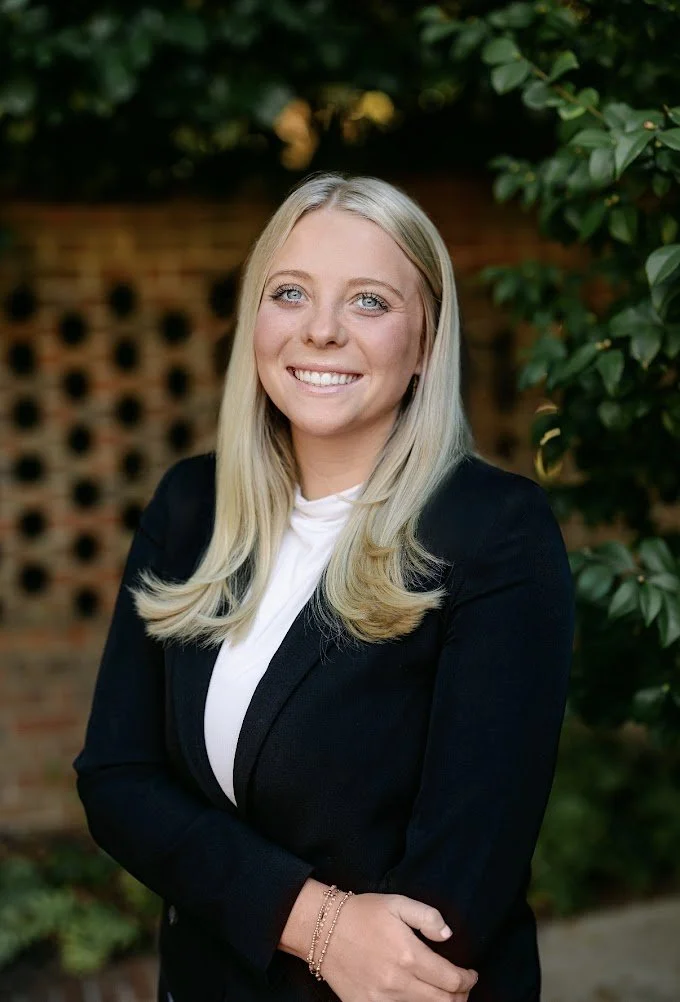 Portrait of a blonde woman with blue eyes, smiling, wearing a black blazer and white top, standing outdoors with greenery and a brick wall in the background.