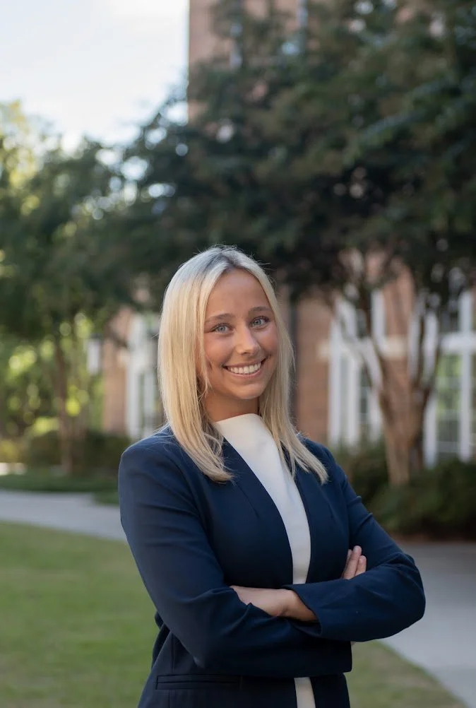A smiling woman with blonde hair wearing a navy blazer over a white shirt, standing outdoors in a park or campus with trees and a brick building in the background.