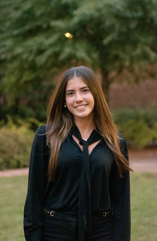 A young woman with long brown hair smiling outdoors in front of green trees, wearing a black blouse with a tie detail and gold accents.