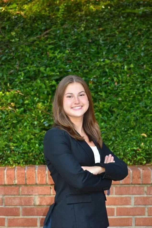 A young woman with brown hair smiling and standing outdoors with arms crossed, in front of a green shrub and brick wall.