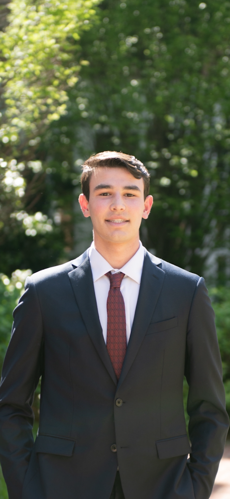 A young man in a dark suit with a white shirt and maroon tie standing outdoors with trees and greenery in the background.