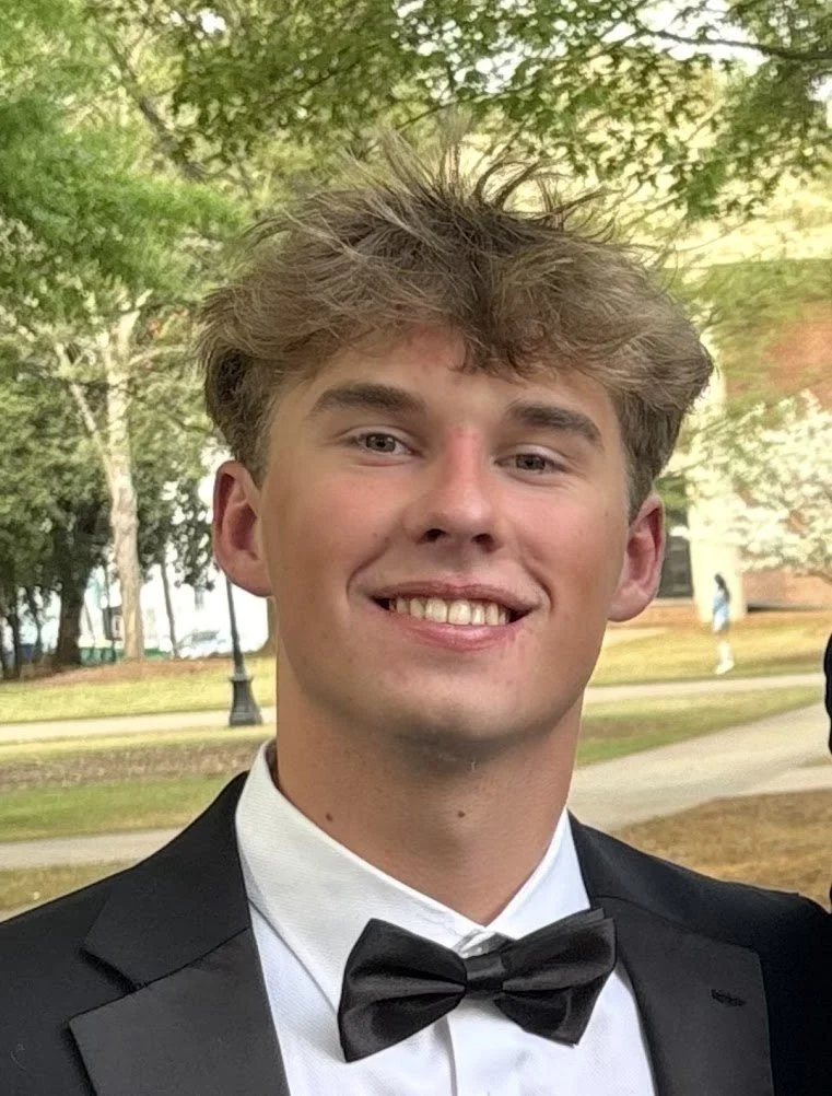 Young man with tousled hair wearing a black tuxedo and bow tie smiling outdoors with trees in the background.