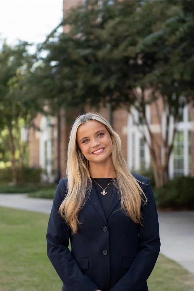 A young woman with long blonde hair smiling outdoors in front of a school building with brick walls and white window frames, dressed in a dark blazer.