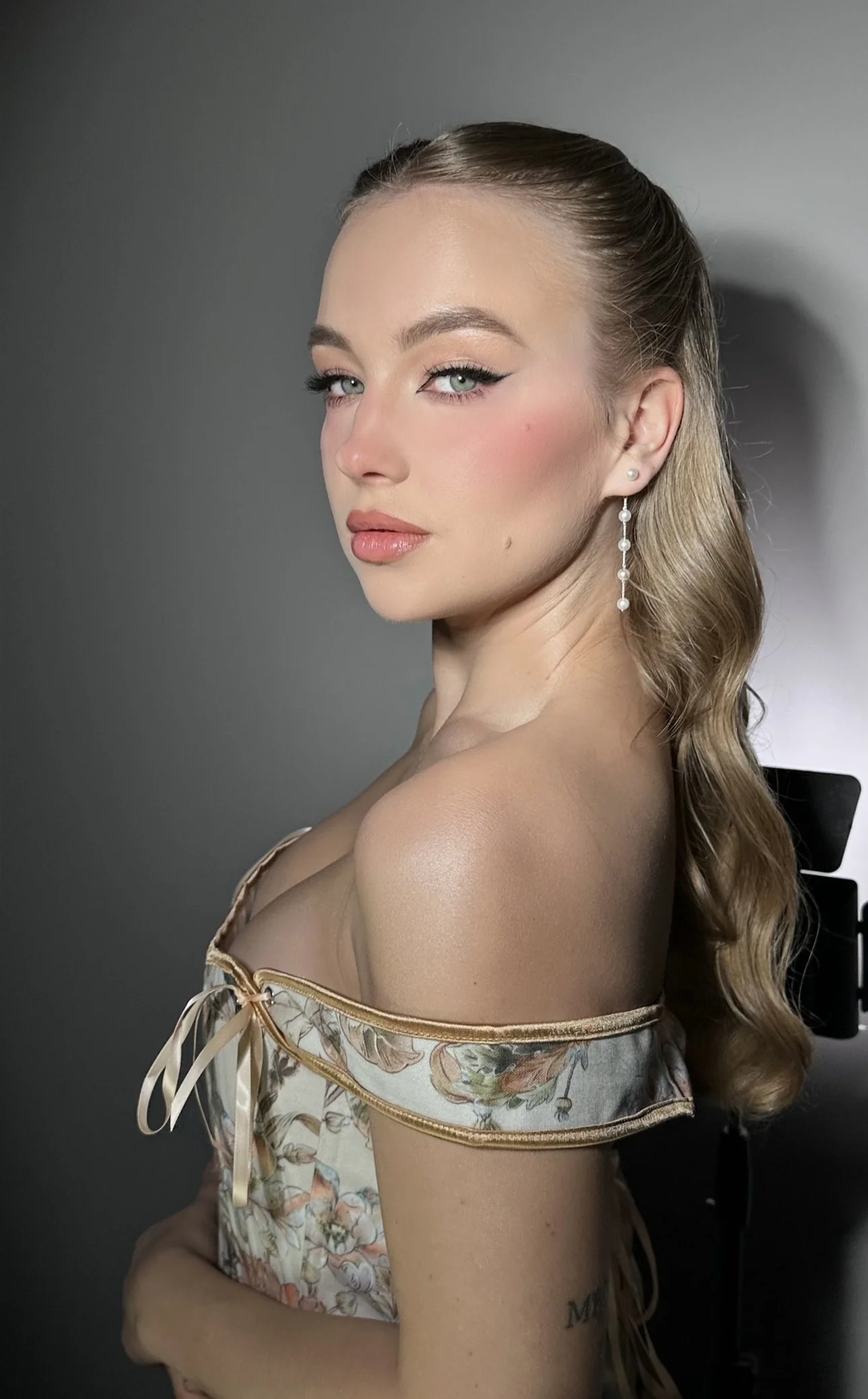 A woman with blonde wavy hair and makeup, wearing pearl drop earrings and an off-the-shoulder floral top, looking at the camera against a plain background.