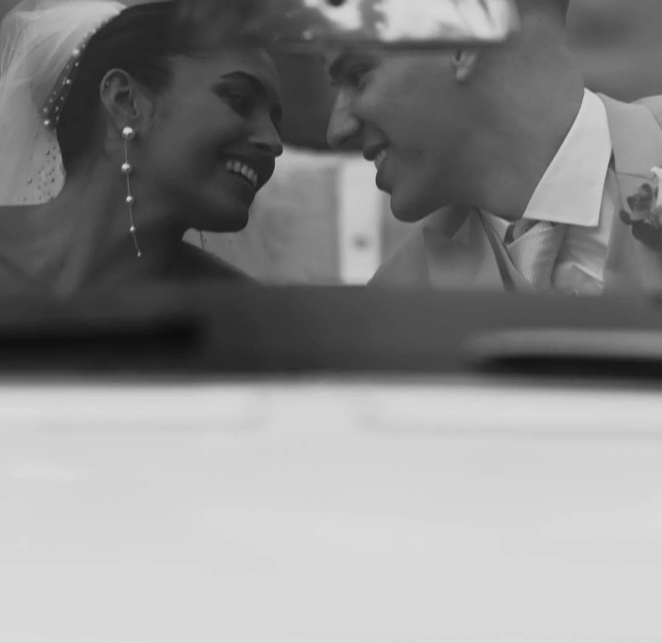 Black and white photo of a bride and groom leaning in close, smiling, with their foreheads touching, visible through a car window.