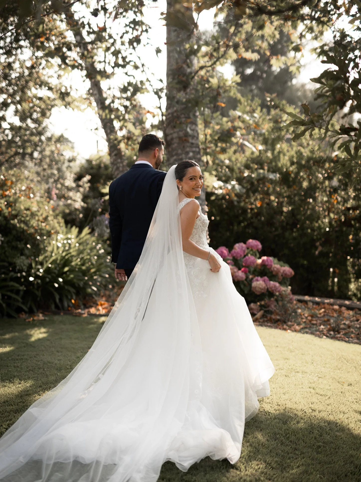 A smiling bride in a white wedding dress with a long veil, posing outdoors with a groom in a dark suit in a garden with trees and pink hydrangea flowers.