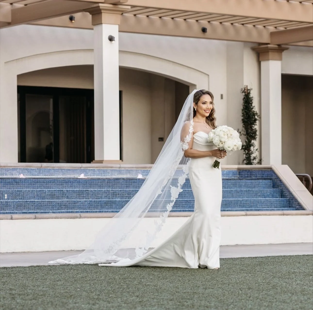 A bride in a white wedding dress holding a bouquet of white flowers stands outside near a swimming pool, with a modern building in the background.