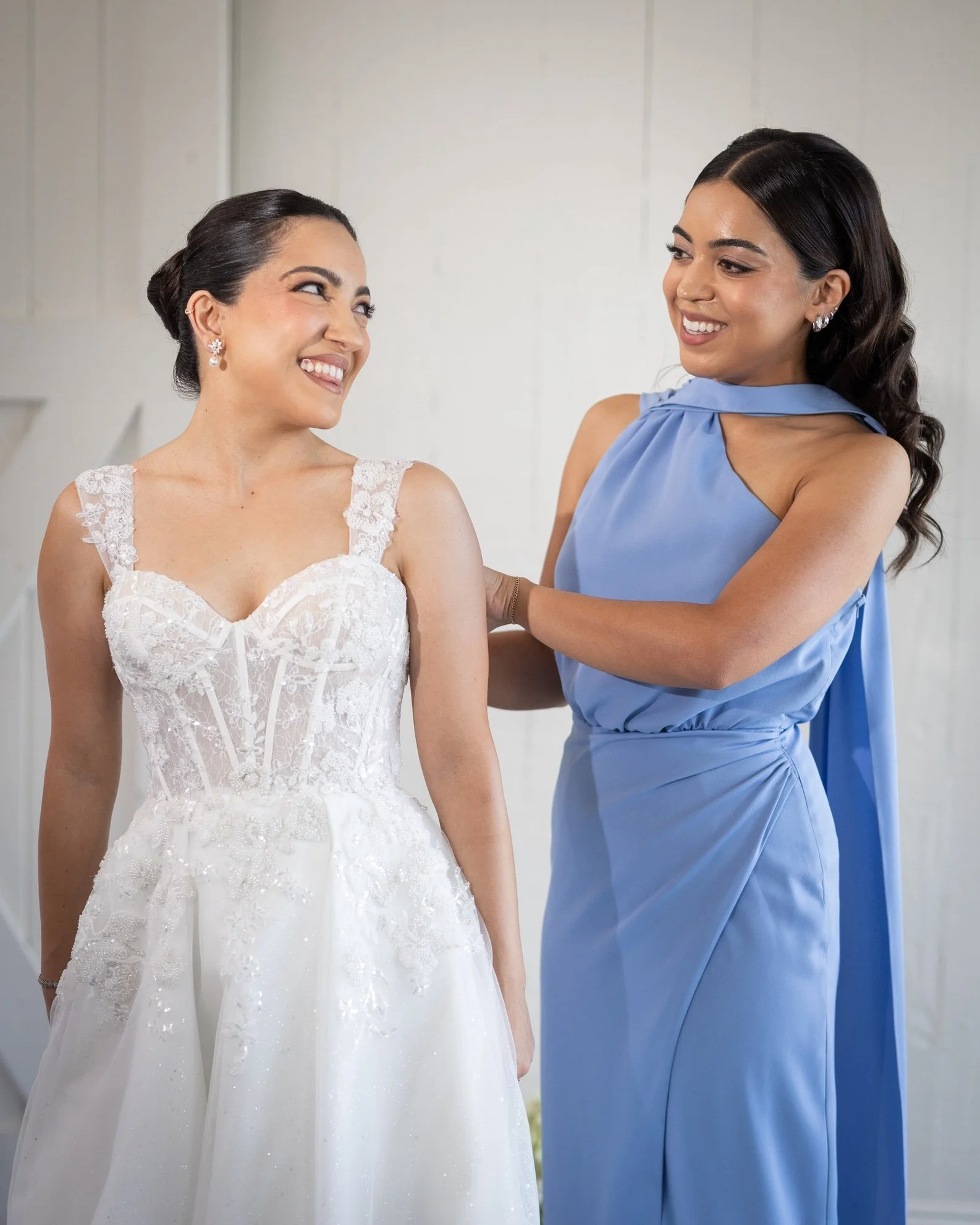 A bride in a white wedding dress smiling at a woman in a blue dress who is laughing, standing indoors against a plain wall.
