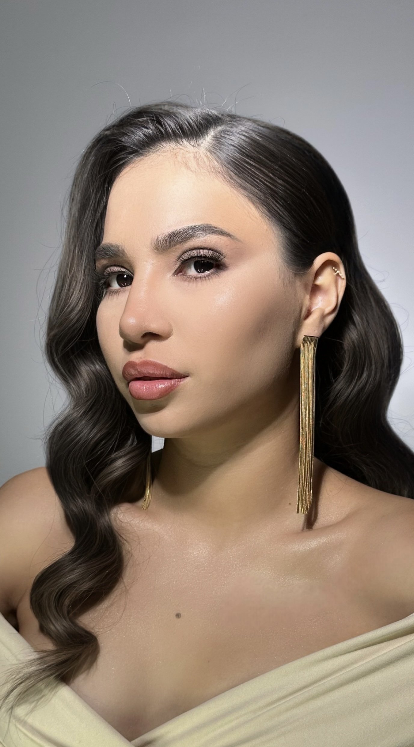 A woman with wavy dark brown hair, wearing long gold earrings, with makeup including defined eyebrows, neutral eyeshadow, eyeliner, mascara, and nude lipstick. She is wearing a beige off-the-shoulder top and has a mole on her collarbone.