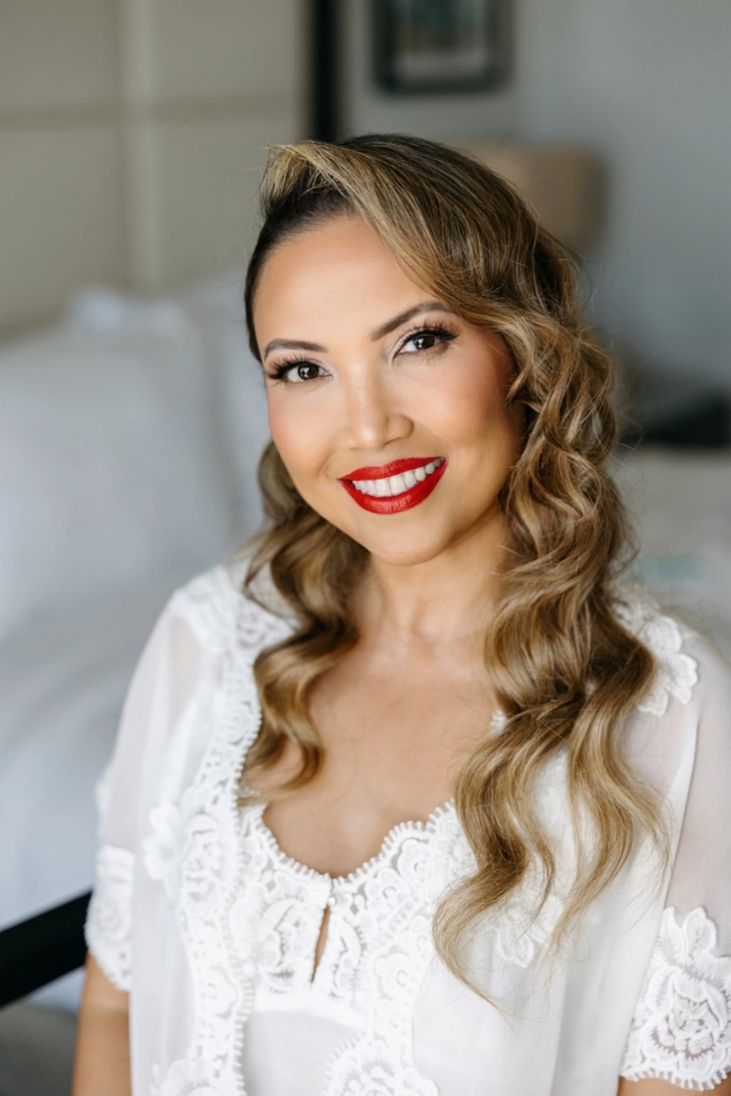 A woman with long wavy hair and red lipstick smiling at the camera in a white lace top, sitting in a bedroom.