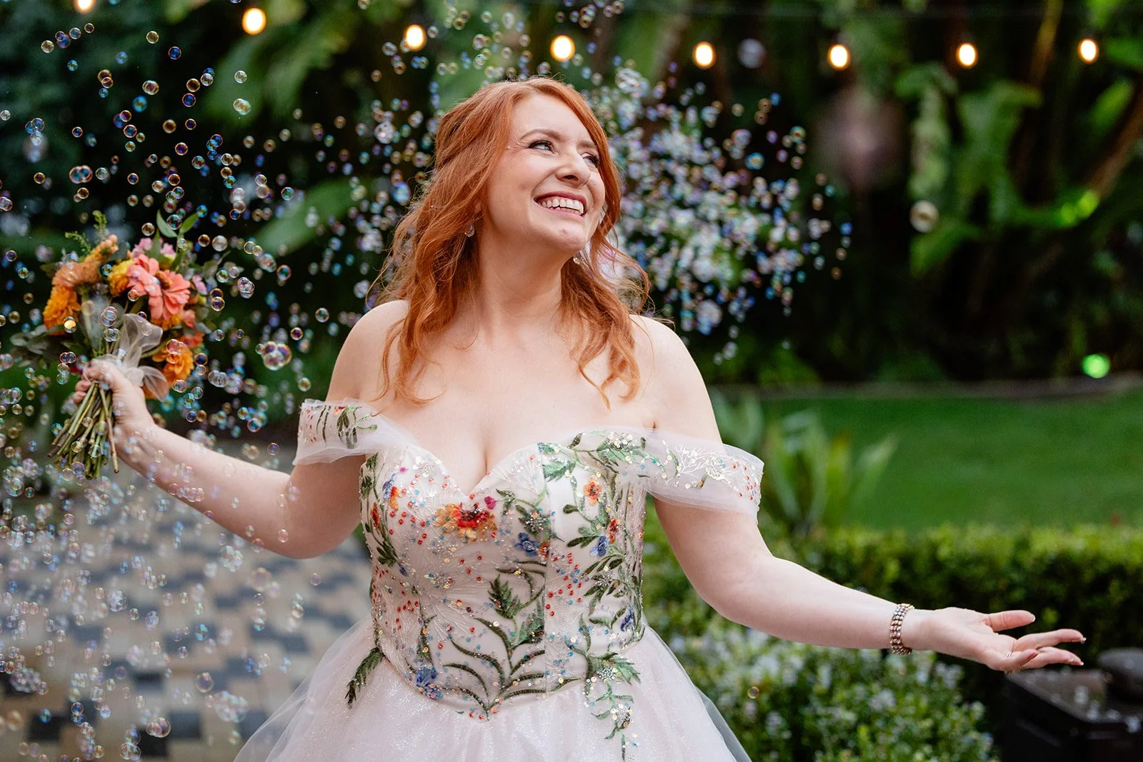 A smiling woman in a floral embroidered wedding dress holding a bouquet, surrounded by floating bubbles in a lush garden setting.
