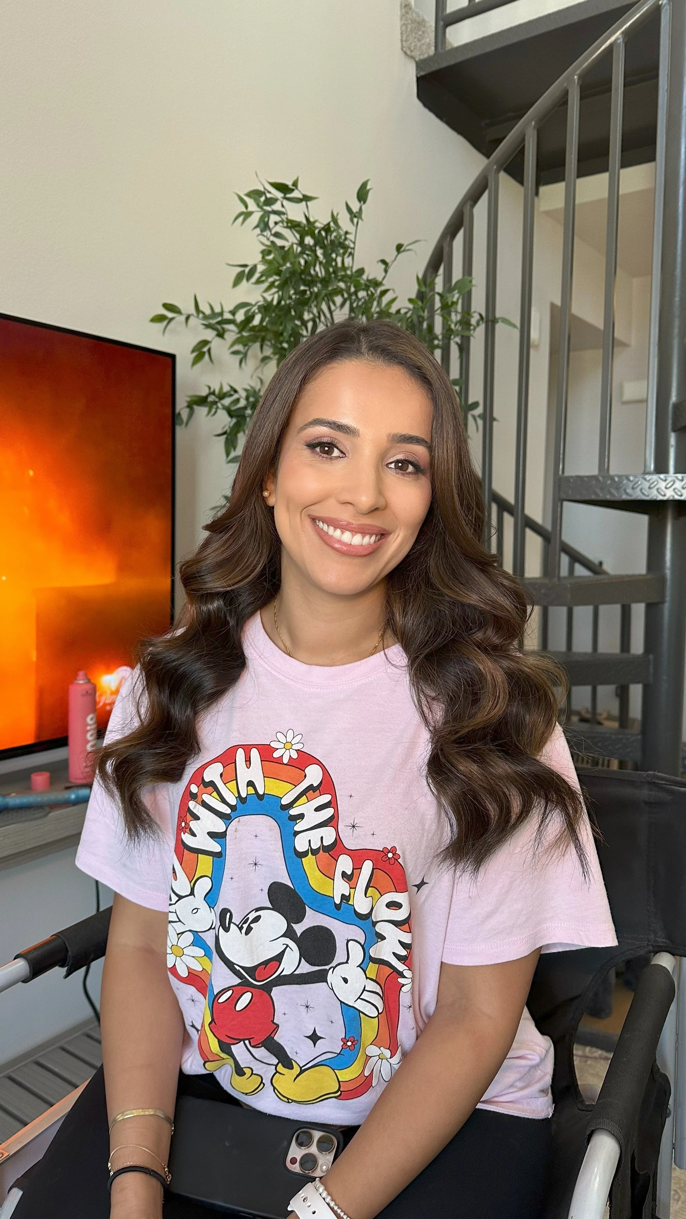 A young woman with long, wavy brown hair smiling at the camera, wearing a pink Mickey Mouse T-shirt with the words "Wish There Was More" and a rainbow design, sitting indoors near a television and a spiral staircase.