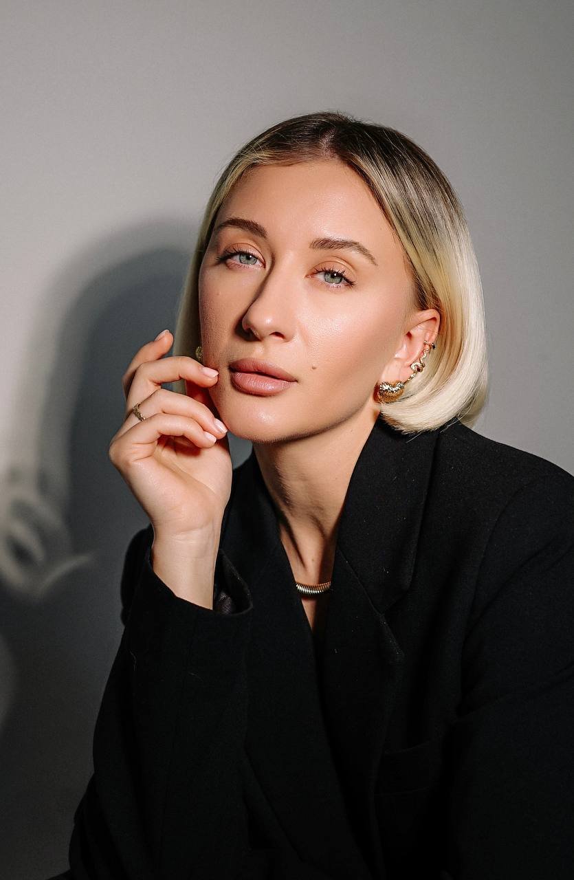 A woman with blonde, shoulder-length hair and light eyes, wearing a black blazer and jewelry including earrings, a ring, and a chain necklace, looking directly at the camera with her hand touching her face against a plain grey background.