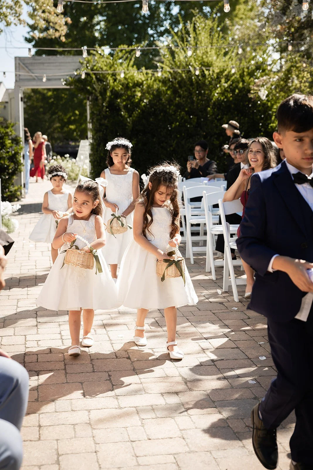 Children dressed in formal attire walking at a wedding ceremony outdoor, with girls in white dresses and boys in suits, on a brick pathway.