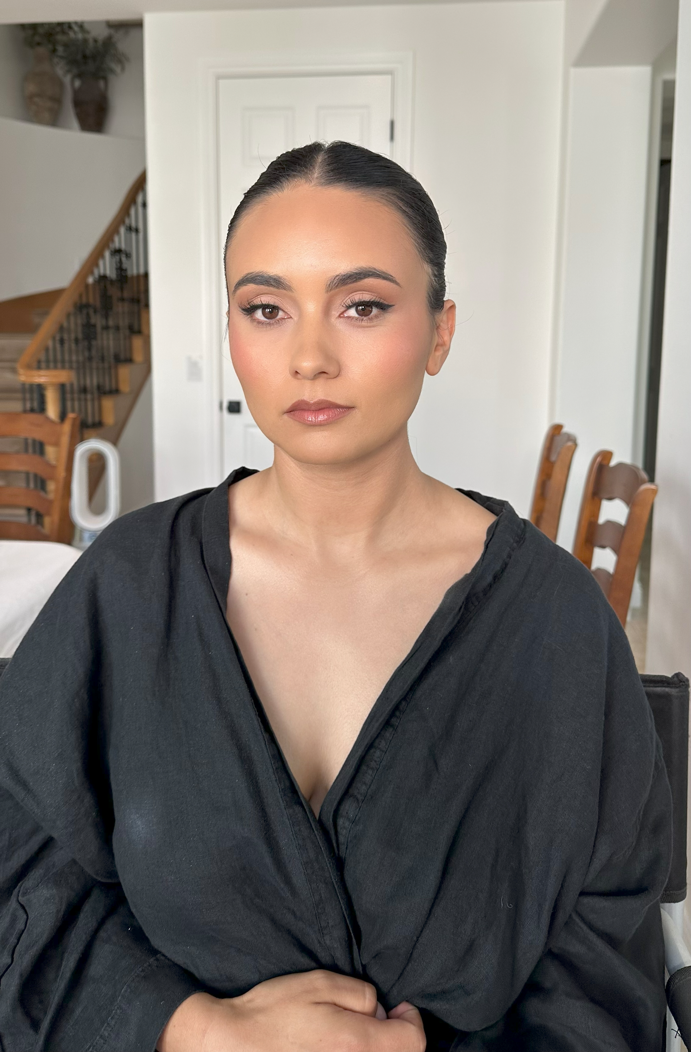 A woman with dark hair pulled back, wearing natural makeup, sitting indoors in front of a white door, dressed in a black top or jacket.