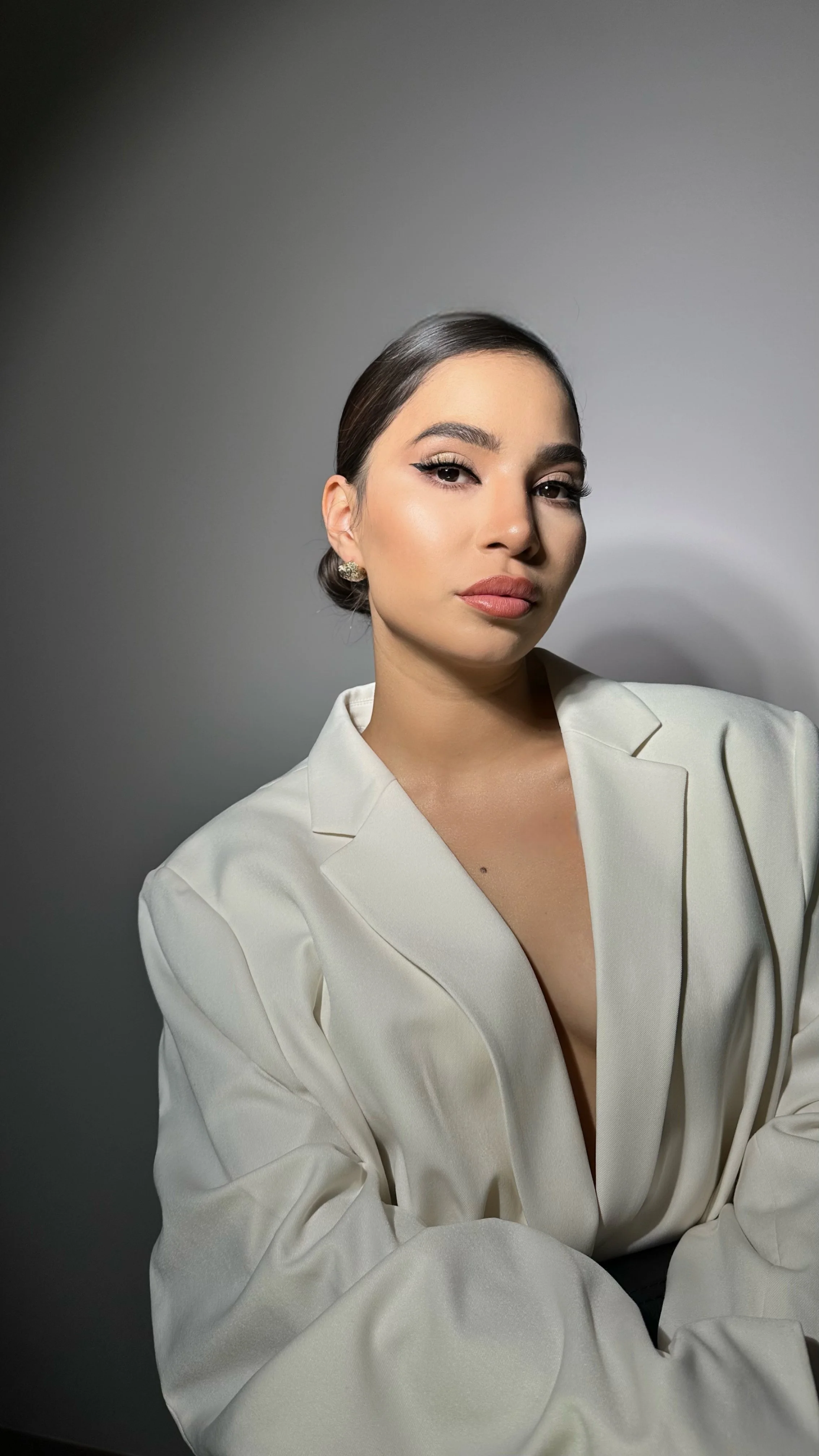 A woman with dark hair pulled back, wearing makeup, earrings, and a cream-colored blazer, posing against a plain gray background.