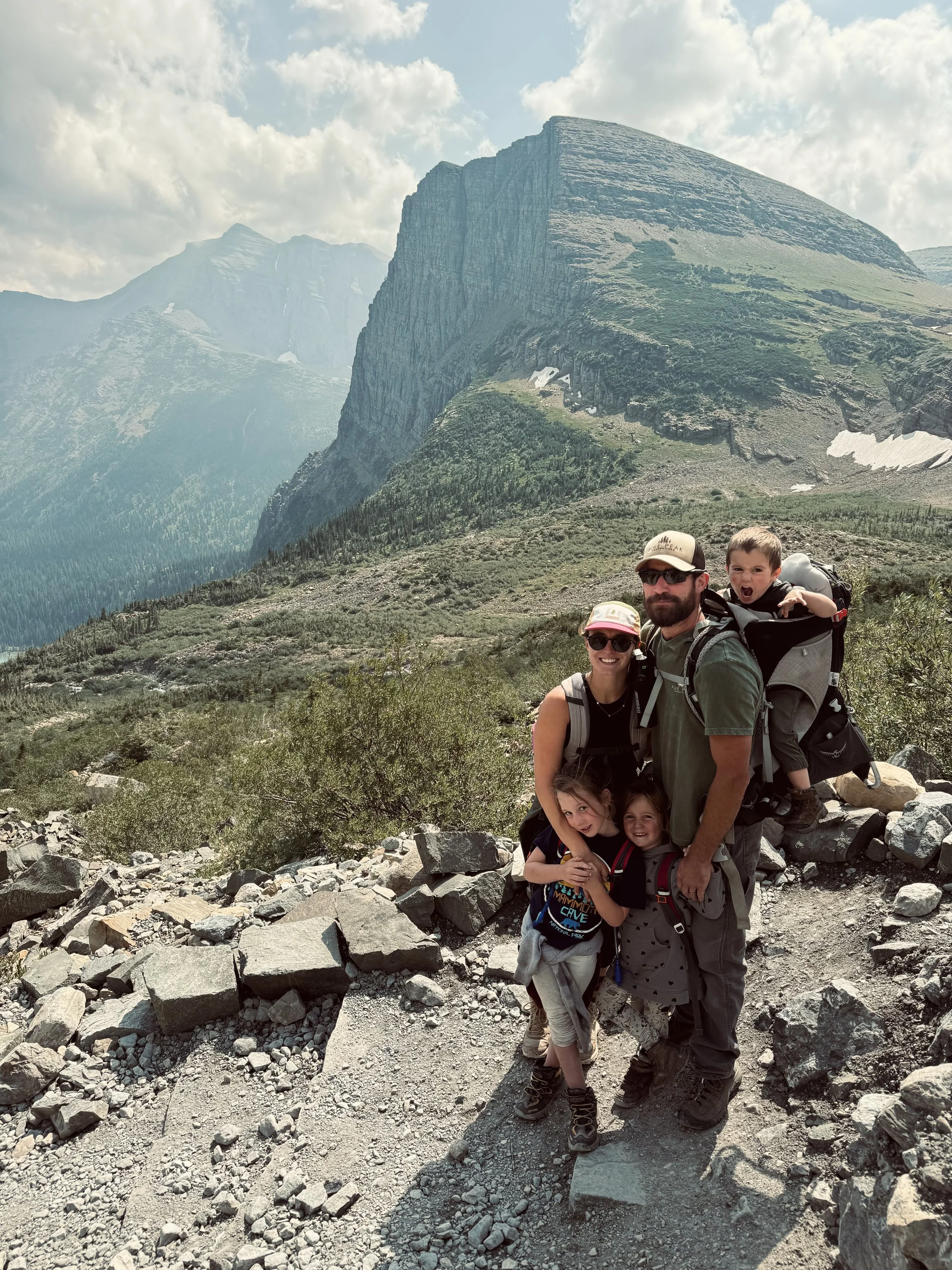 Family hiking in a mountainous area with rocky terrain, green bushes, and large cliffs in the background.