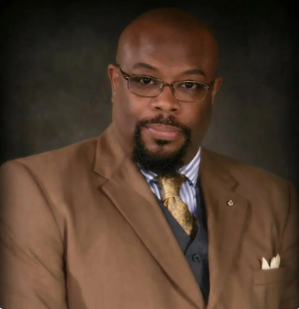 Professional portrait of a bald African American man with glasses, dressed in a brown suit jacket, blue striped shirt, and gold tie, against a dark background.