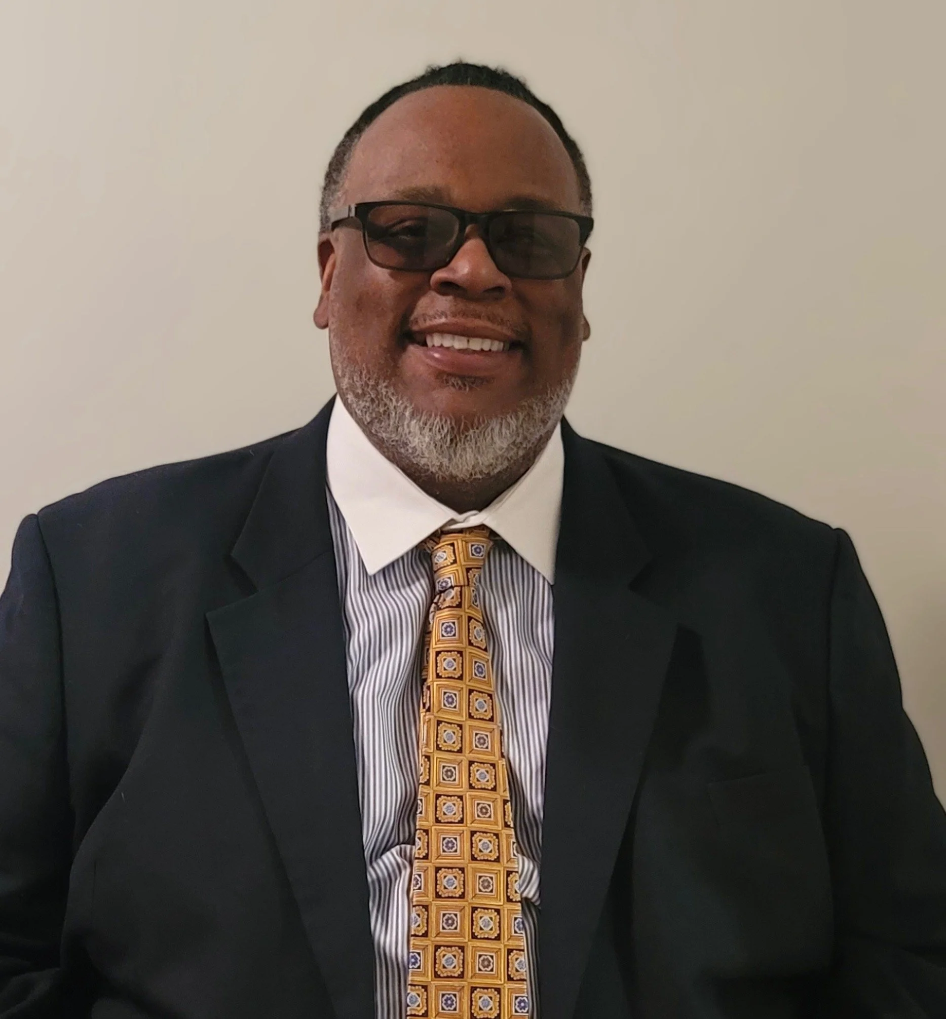 A smiling middle-aged man wearing glasses, a dark suit, a striped shirt, and a yellow patterned tie, sitting against a plain light-colored wall.