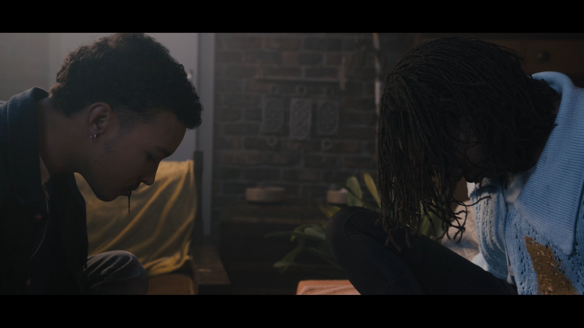 Two people sitting on the floor with heads bowed, in a dimly lit room with a brick wall in the background.