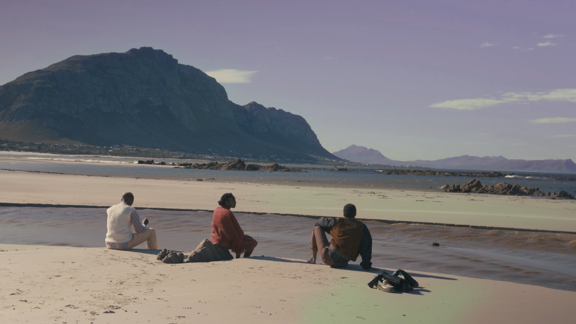 Three people sitting on a sandy beach facing the ocean with mountains in the background.