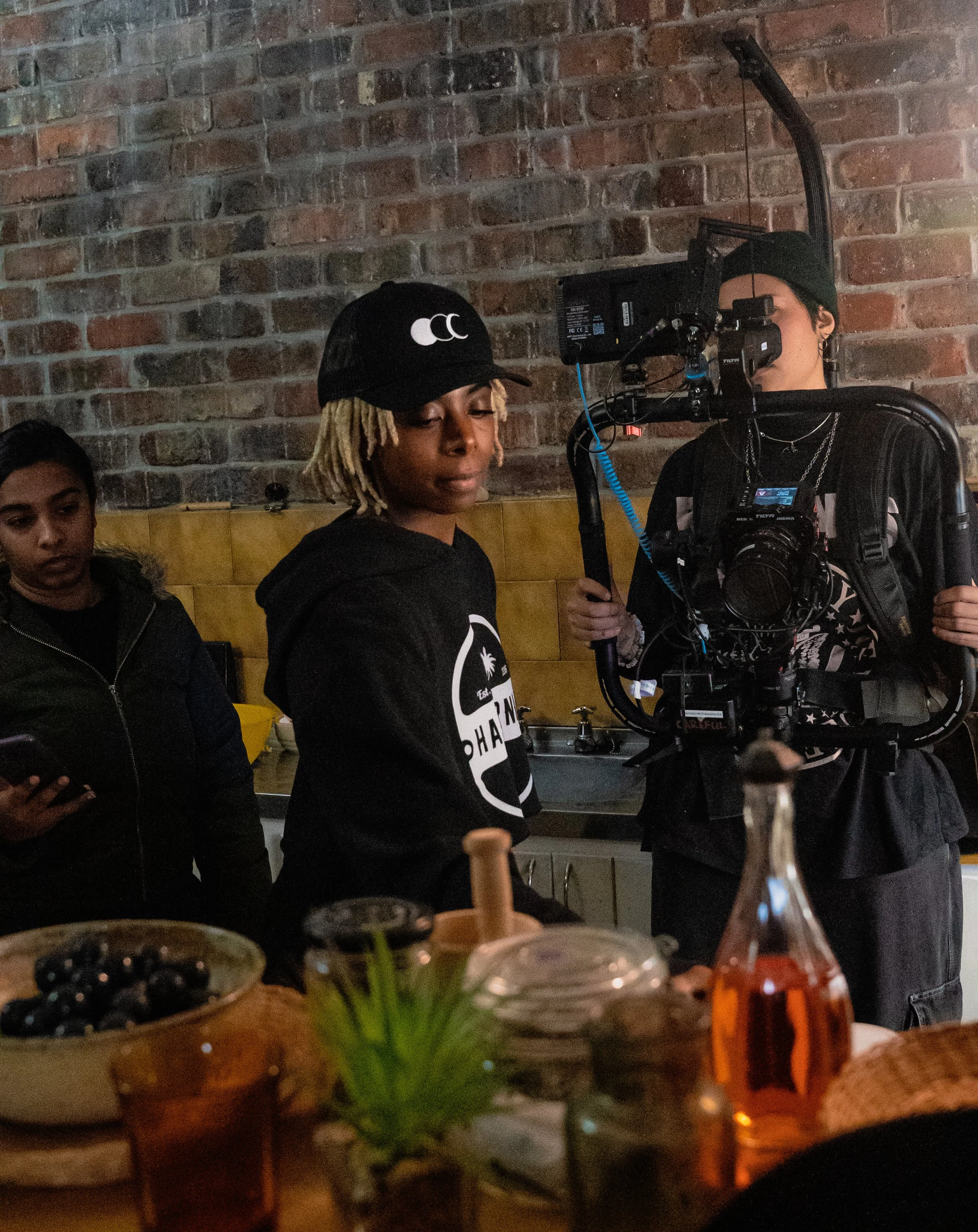A person with blonde dreadlocks and a black cap filming a woman with dreadlocks in a black hoodie, inside a restaurant with exposed brick walls. Another woman is visible on the left side of the image, looking at her phone.