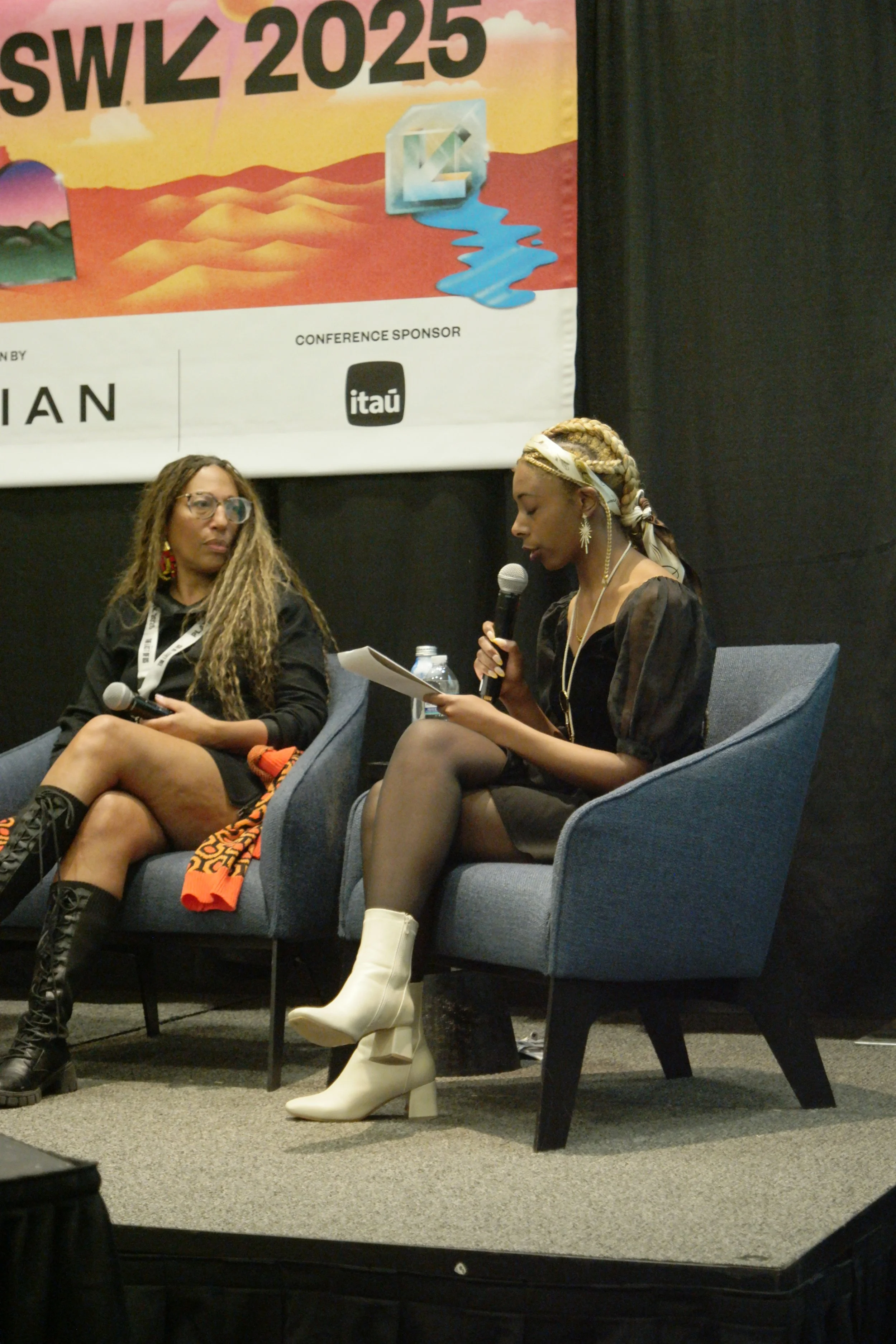 Two women sitting on stage chairs, one speaking into a microphone and reading from notes, at a conference with a large colorful banner behind them.
