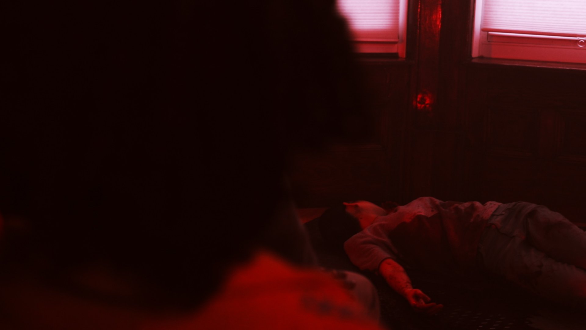 A person lying on the floor in a dimly lit room with red lighting, with a window behind them and another person's head partially visible in the foreground.