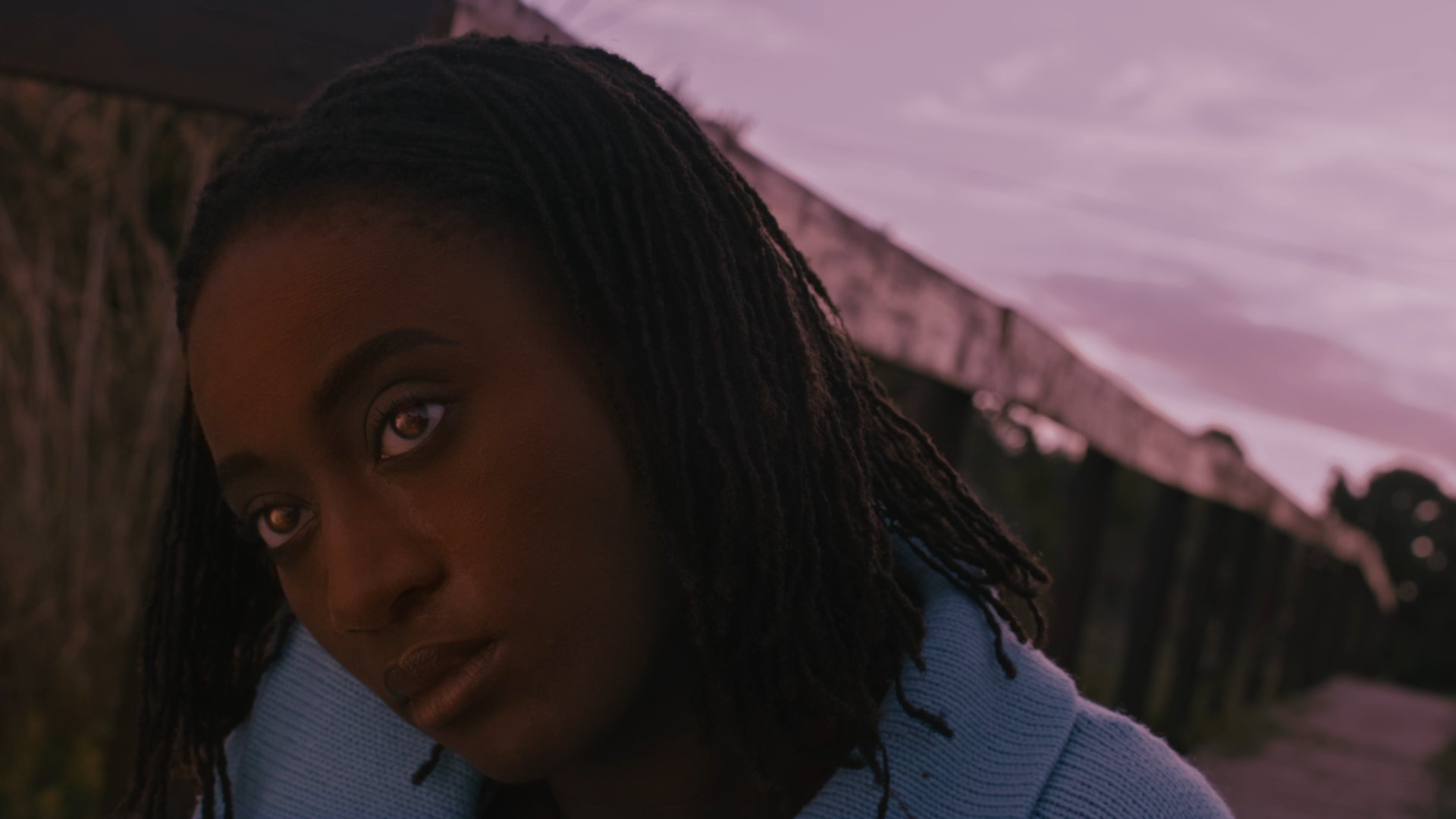 Close-up of a young woman with braided hair looking into the camera with a serious expression, outdoors during sunset with a purple sky.