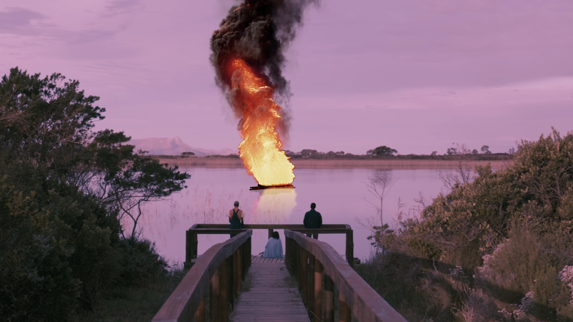 A boat is on fire in a body of water with large flames and black smoke, seen from a wooden dock where three people are watching at sunset or dawn with purple sky.
