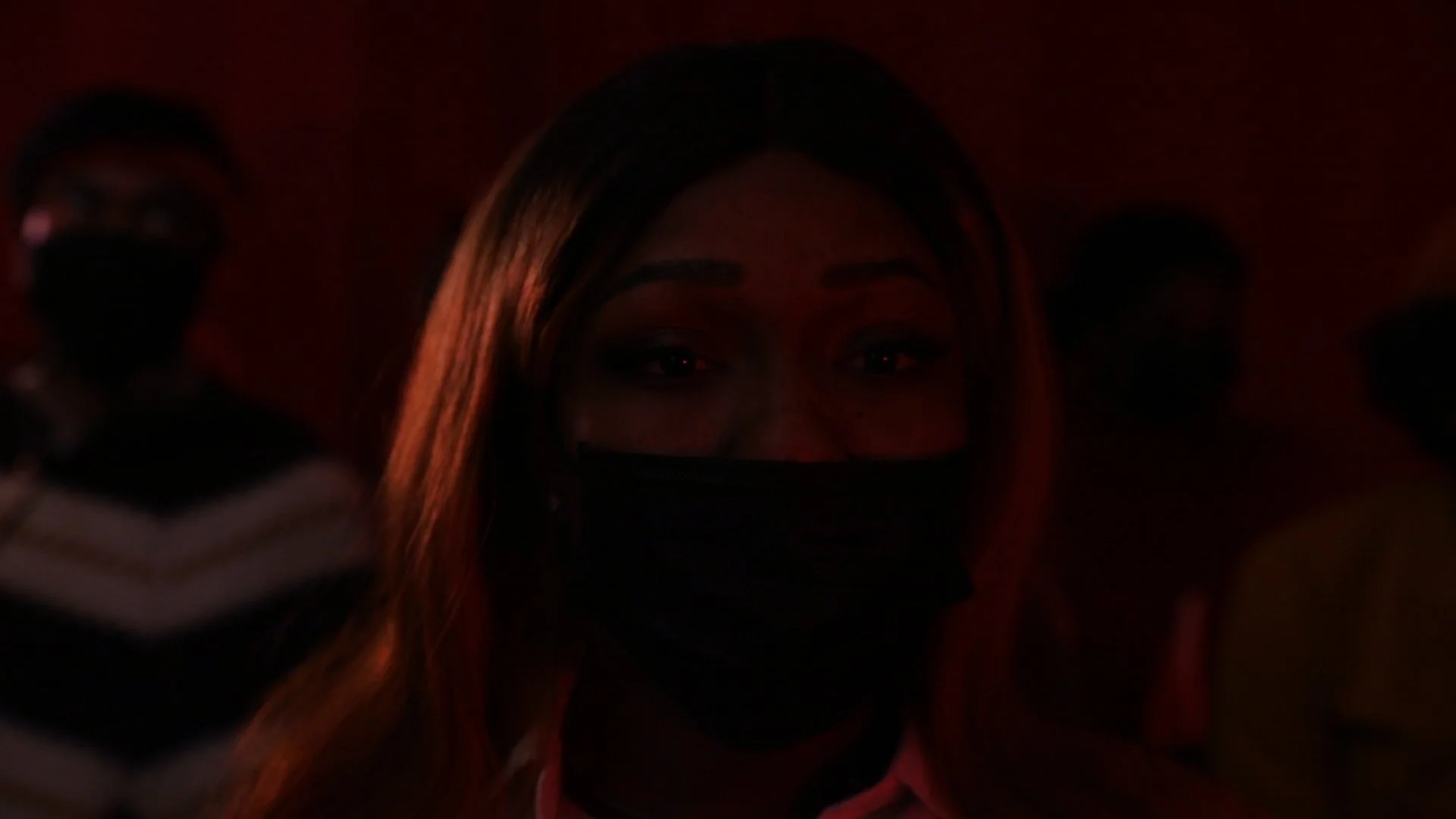 A woman with long hair wearing a black face mask, looking directly at the camera in a dimly lit environment with reddish lighting. Two other people are partially visible in the background.