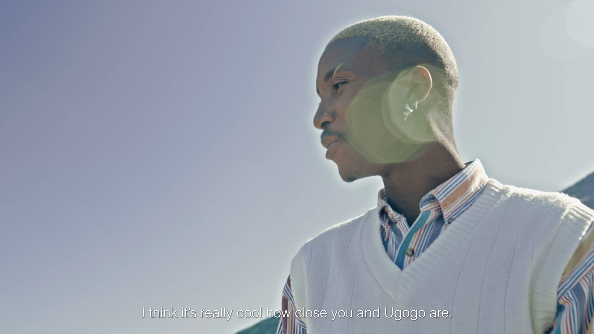 A young man with short blonde hair, earrings, wearing a striped shirt and a white vest, stands outdoors against a clear sky, talking to someone off-screen.