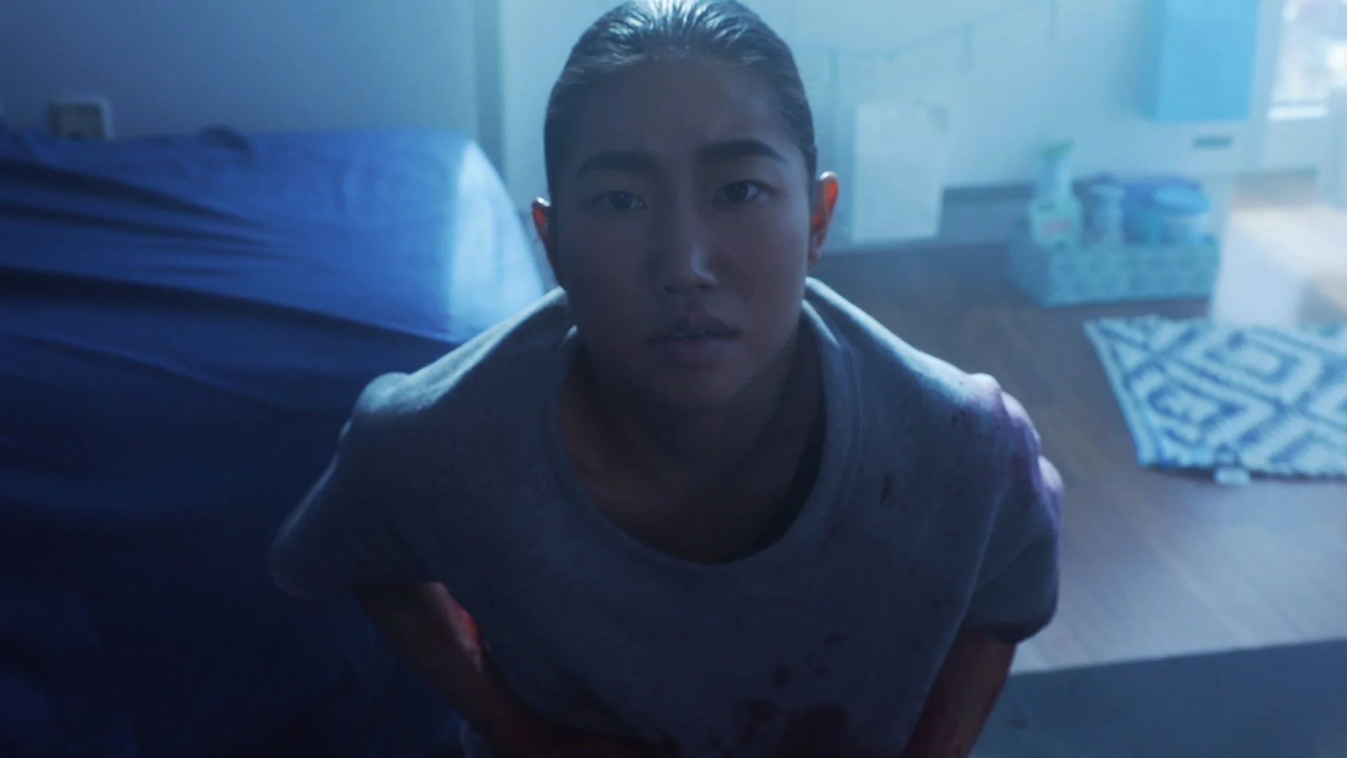 A young man with slicked-back hair looking directly at the camera inside a classroom or medical setting, with some supplies visible in the background.