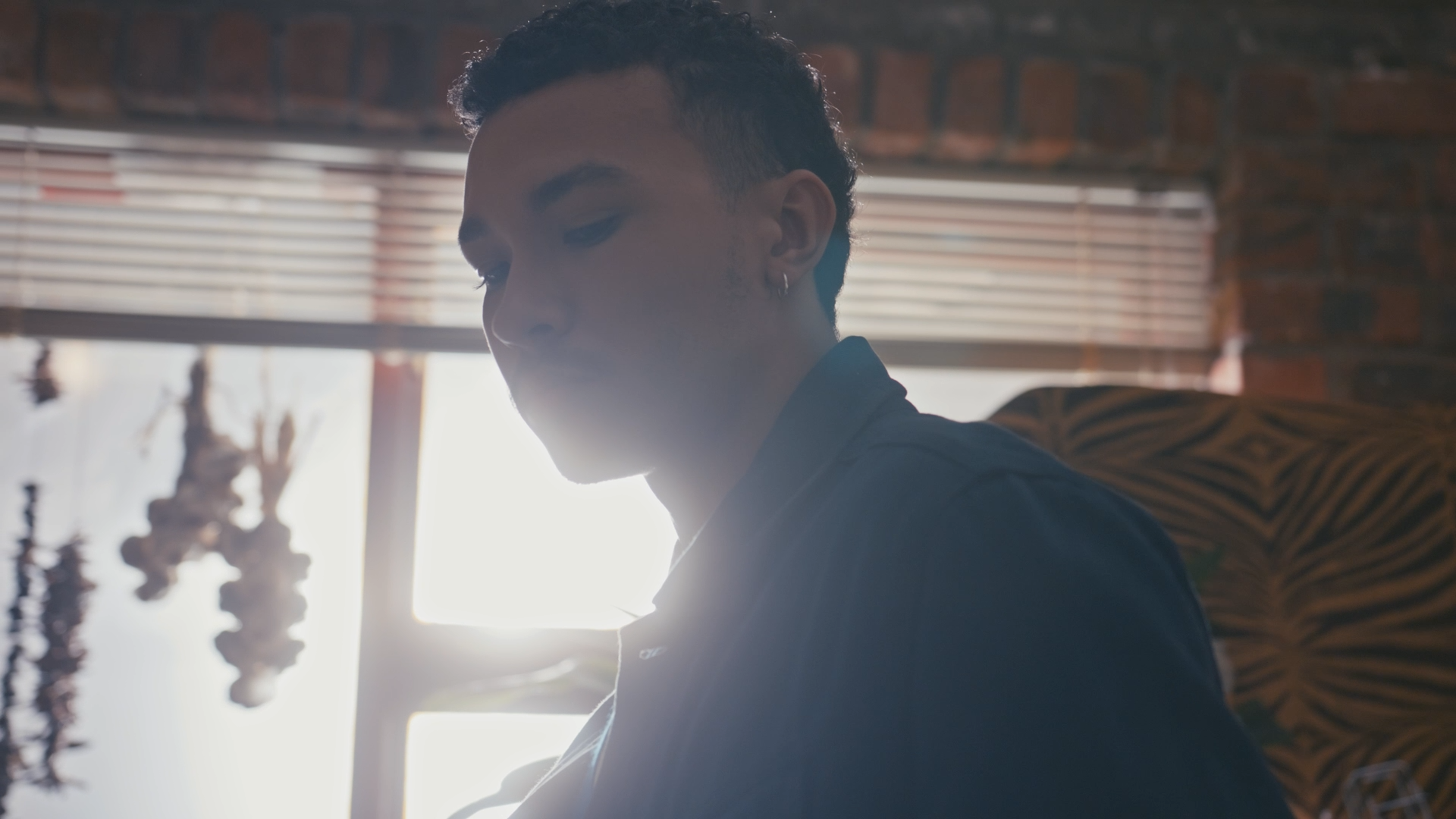 A young man with earrings looking down, sitting near a window with blinds, and a brick wall behind him, backlit by sunlight.