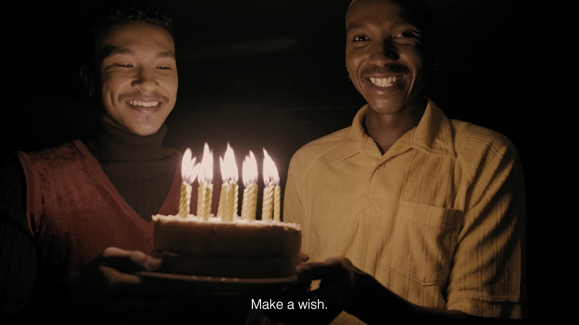 Two people, one in a dark turtleneck and vest and the other in a yellow shirt, smiling and holding a birthday cake with lit candles in a dark room.
