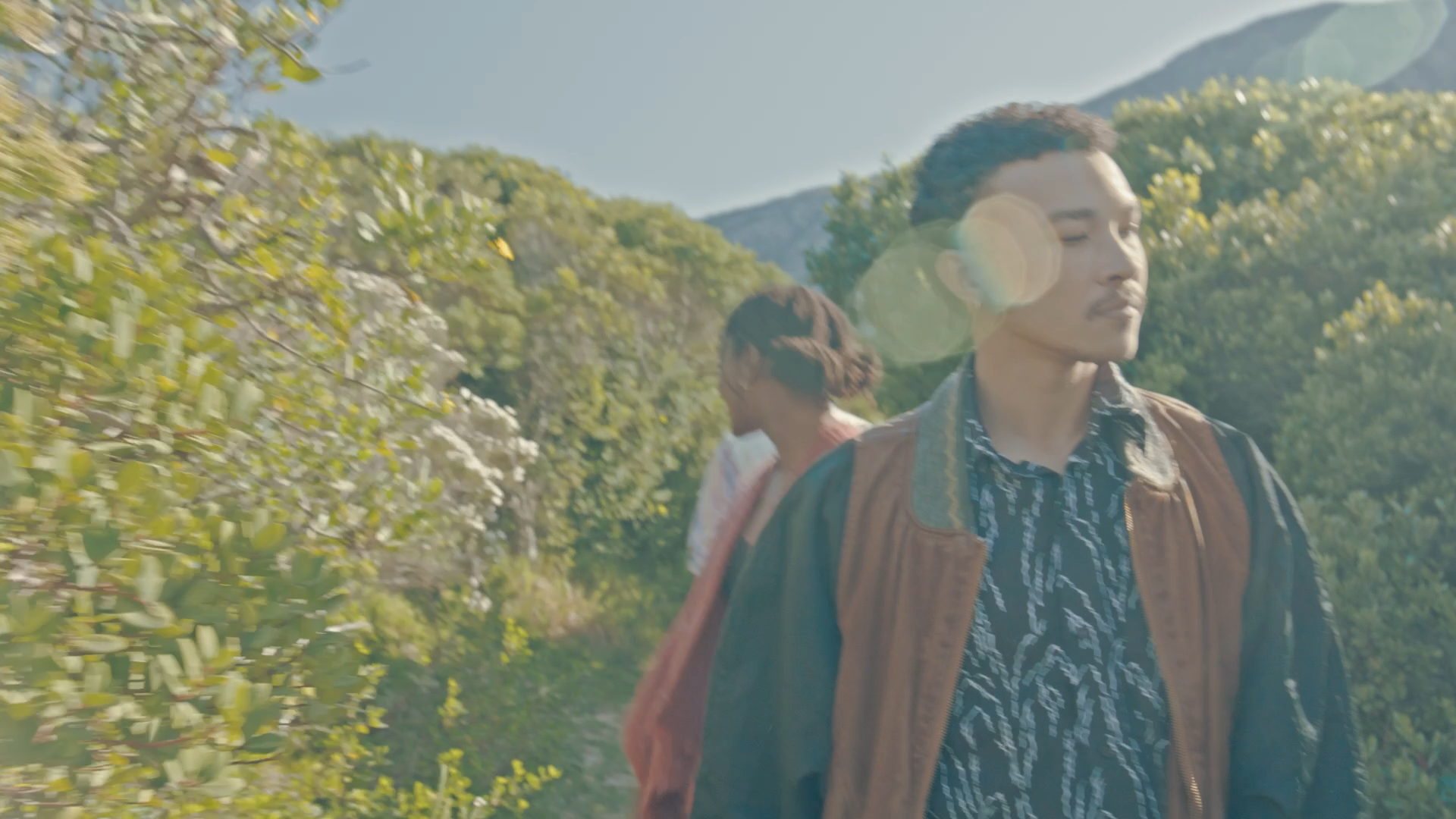 A man and a woman walking outdoors in a green, mountainous area with sunlight flares.