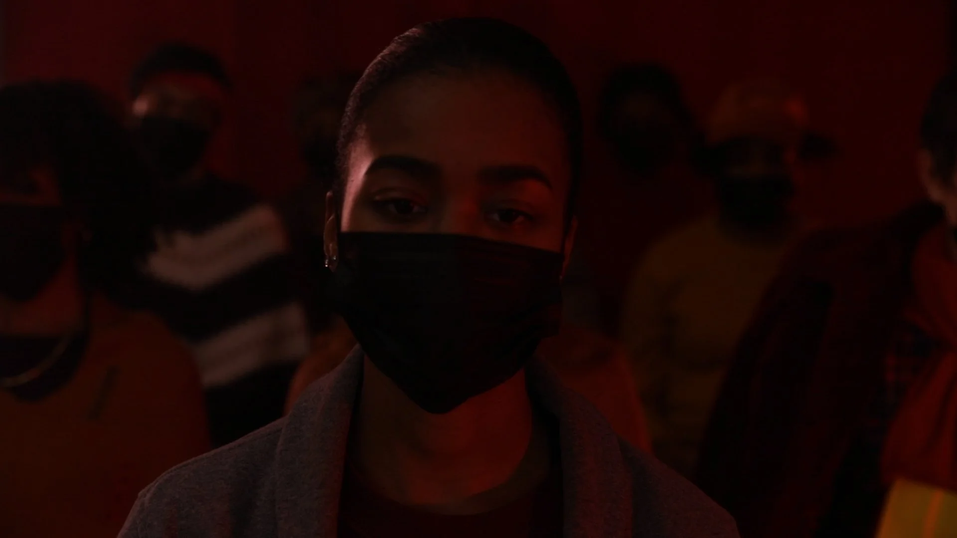 Person wearing a black face mask in a dimly lit indoor setting with other people in the background.