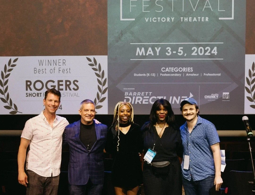 Five people posing on stage at an award ceremony with a large screen behind them displaying event information, including winners and categories for a film festival.