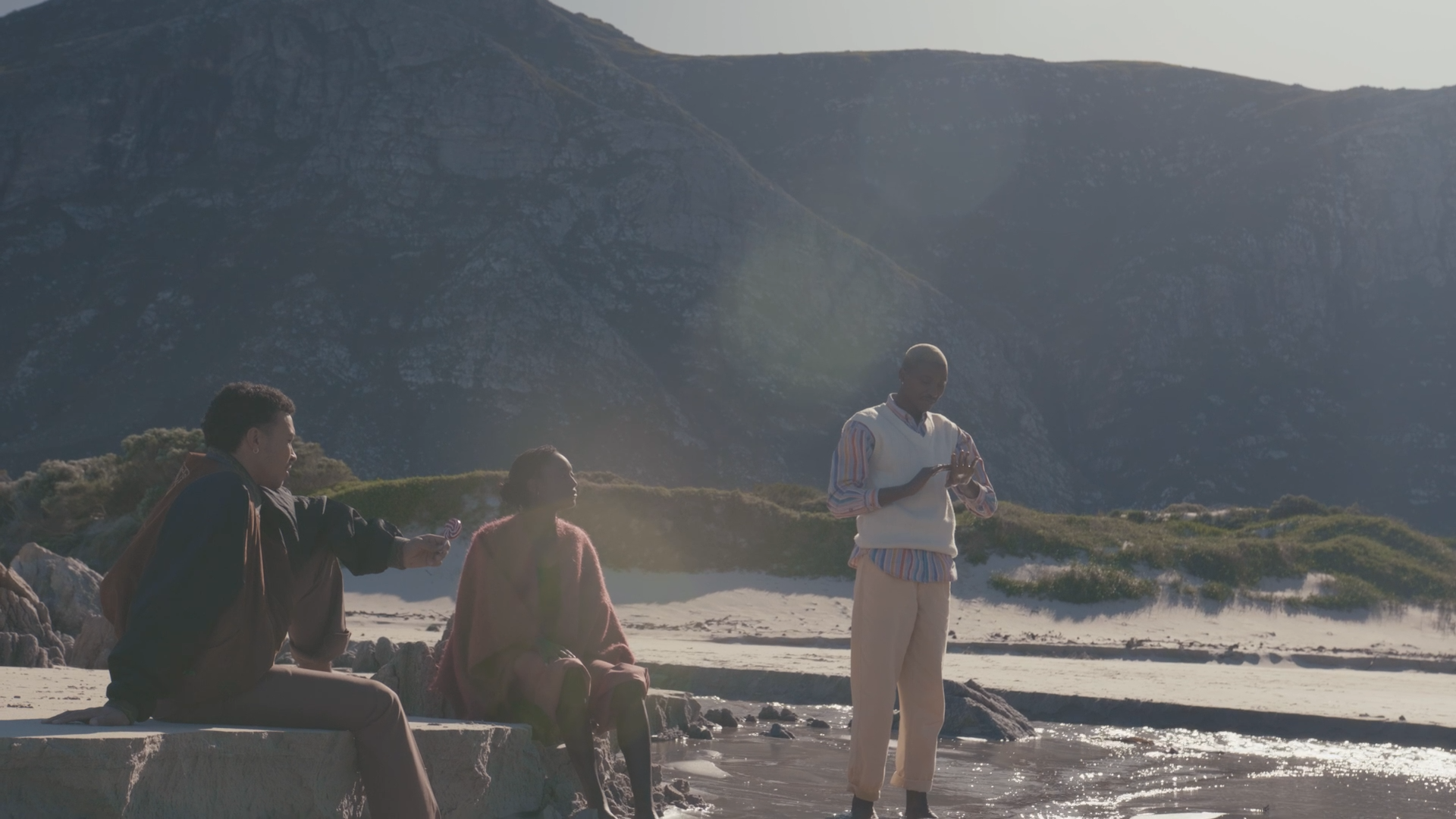 Four young people at the beach with mountains in the background, one sitting on a ledge and the others standing or sitting on the sand, with sunlight and water reflections.