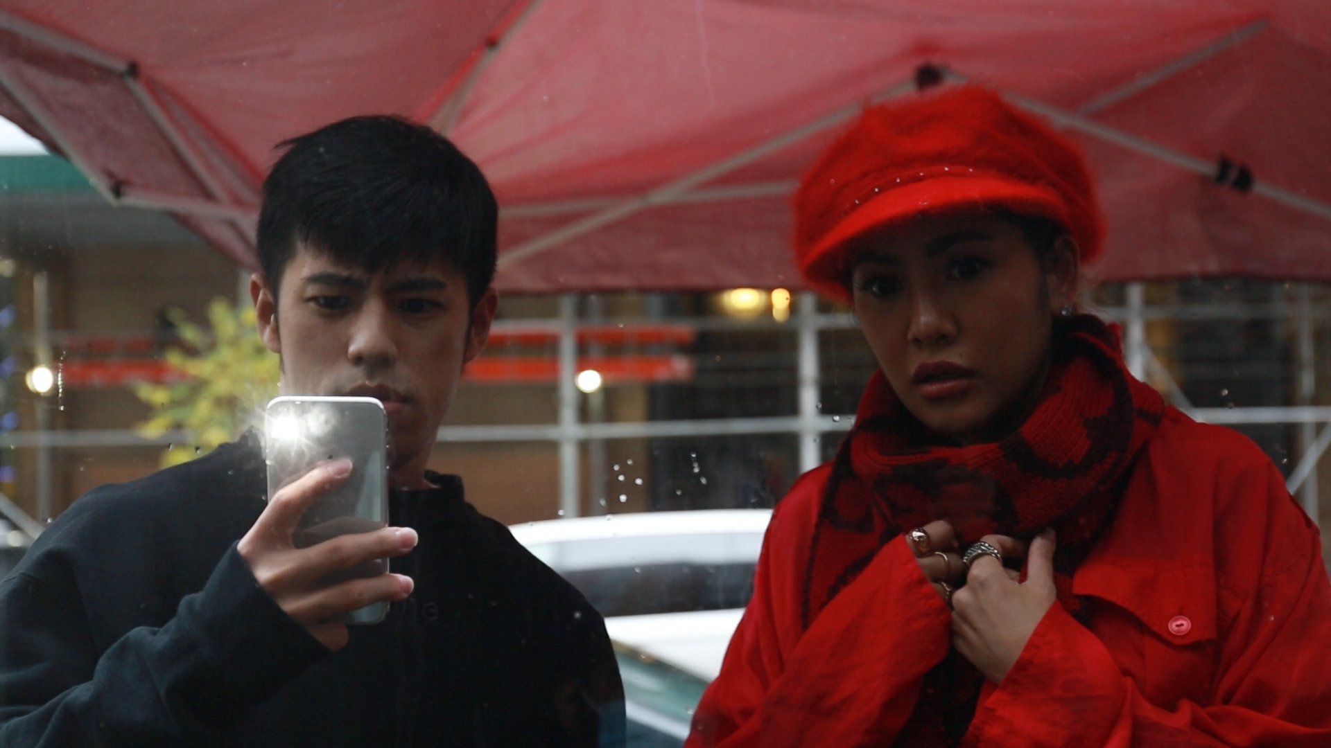 A man and a woman standing outside in the rain under a red umbrella, looking at a phone. The man is holding the phone and wearing a black jacket. The woman is dressed in red, wearing a scarf, hat, and jacket, and looks surprised or concerned.