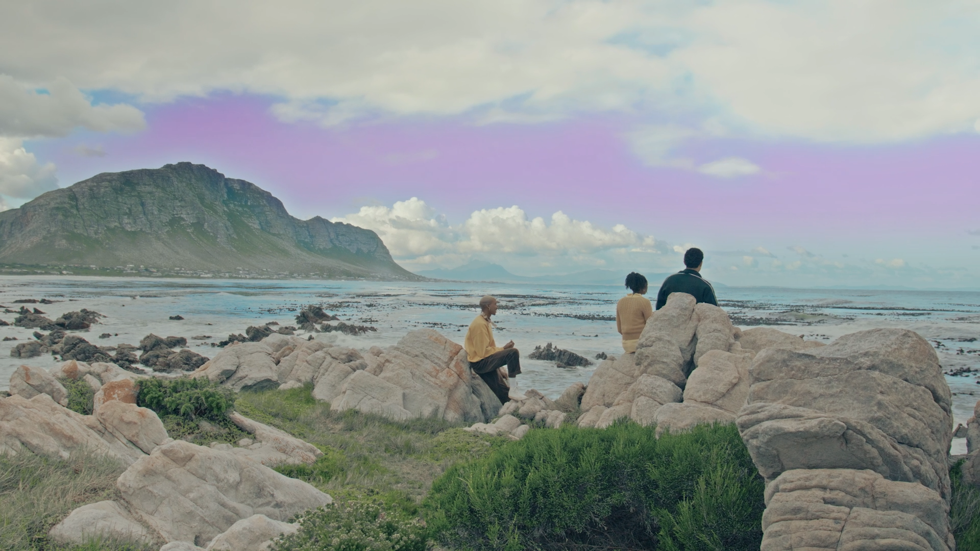 Three people sitting on rocks by the ocean with mountains in the background and a cloudy sky.