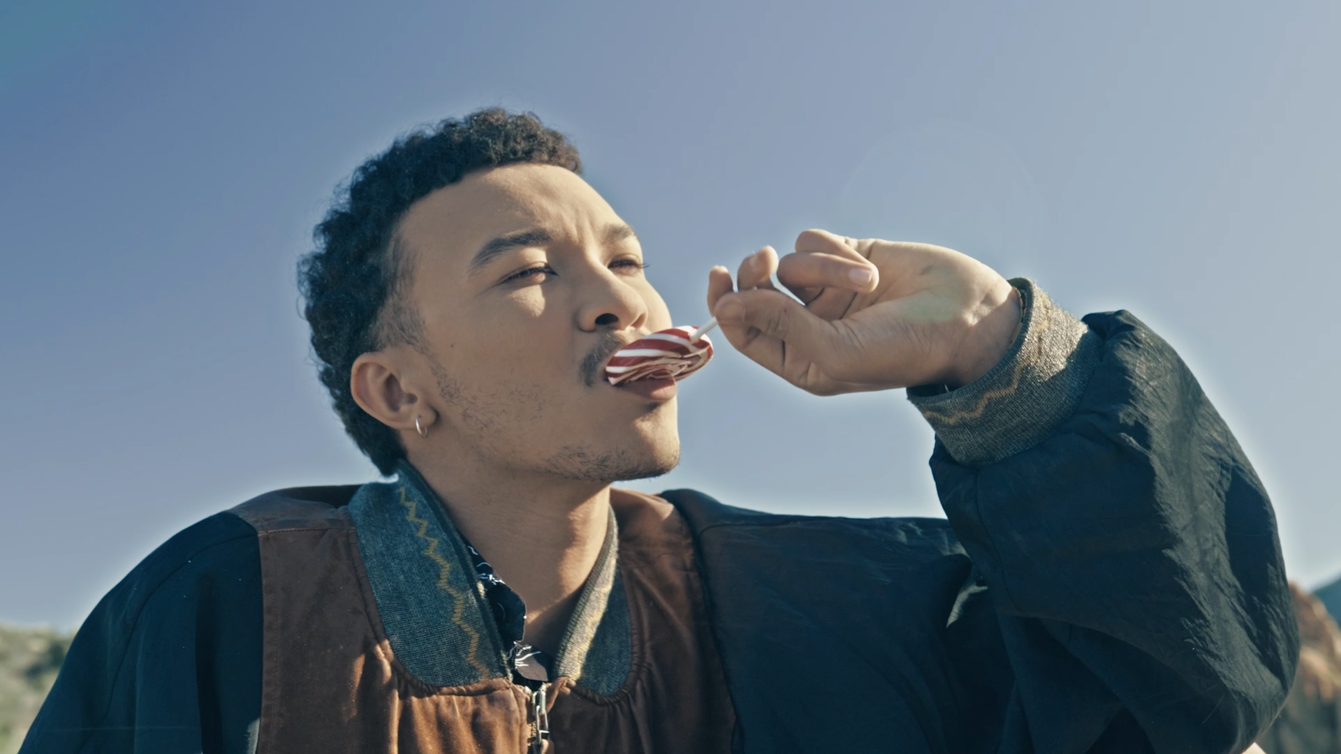 A man with dark hair and a mustache licks a red and white striped lollipop outside under a clear blue sky.
