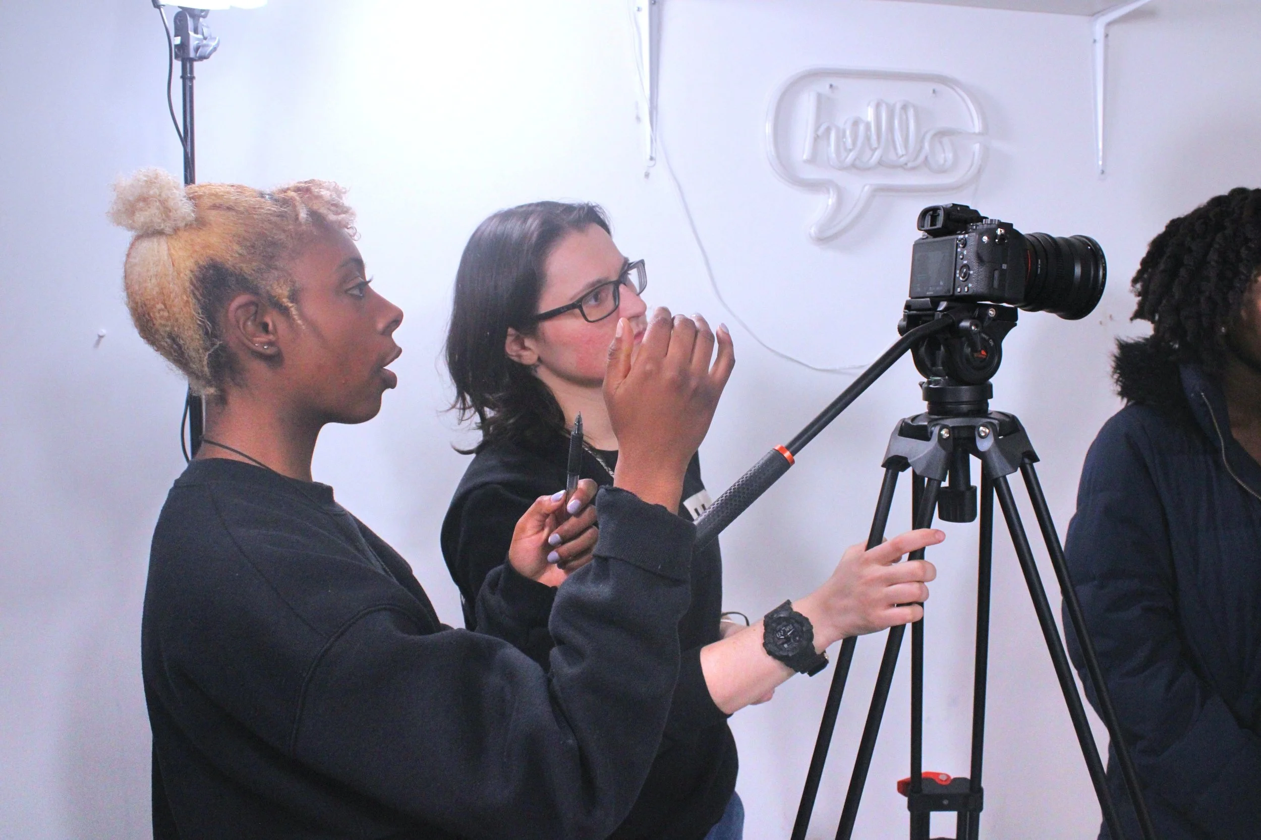 Two women outdoors at night, one with dyed blonde hair and the other with glasses, looking at a camera on a tripod, with a neon sign in the background.